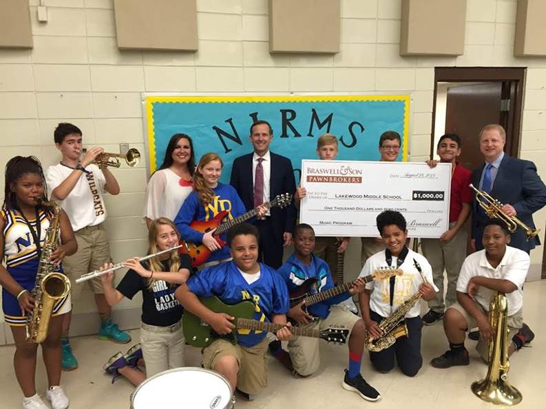 Douglas Braswelll of Braswell & Son Pawnbrokers in Little Rock, Arkansas with large donation check and school faculty, staff and students holding donated musical instruments.